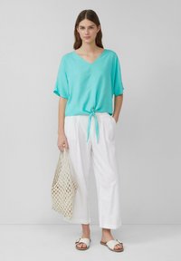 Top turchese a maniche corte con scollo a V e nodo frontale; abbinato a pantaloni bianchi a gamba larga e sandali. Tiene una borsa a rete beige.