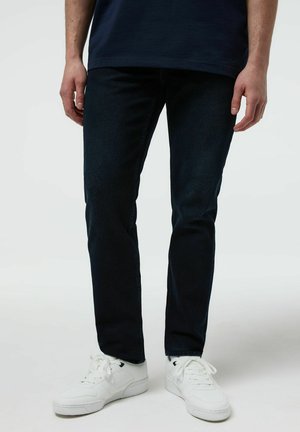 Mężczyzna ubrany w ciemne, dopasowane jeansy i białe trampki, stoi z rękami swobodnie opuszczonymi przy bokach na jednolitym tle.