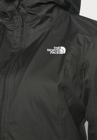 Zwarte capuchonjas gemaakt van een gladde, lichte stof. Beschikt over een voorrits en een wit logo op de linker borstzijde.