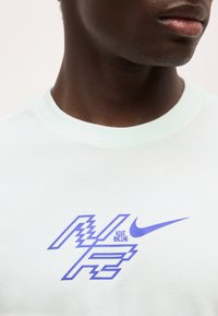 Κοντινή λήψη ατόμου που φοράει λευκό πουκάμισο με μπλε λογότυπο Nike Racing και το σήμα swoosh στο στήθος.