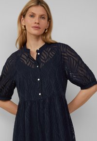 Robe bleu marine avec motif texturé en vagues, manches courtes, boutons sur le devant et taille froncée. Tissu transparent renforce l'effet de superposition.