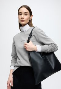 Nagy fekete bőr válltáska sima felülettel és egy vállpánttal, szürke pulóverrel és fehér garbóval párosítva.