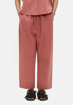 Kobieta nosząca luźne, wysokowalne spodnie w kolorze przygaszonego różu z troczkiem oraz dopasowaną, zapinaną na guziki koszulę, zestawione z czarnymi, odkrytymi sandałami z frędzlami.