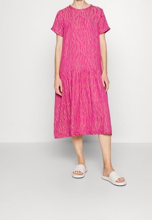 Kvinde iført en midi-kjole med pink og mauve mønster, korte ærmer og hvide slid-sandaler står på en ensfarvet baggrund.