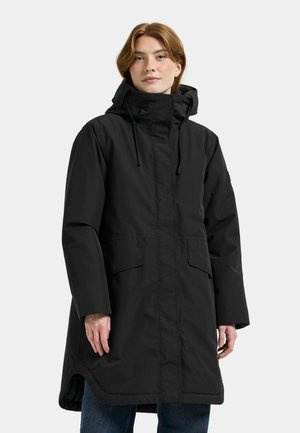 Kvinde iført en lang sort vinterjakke med hætte, store frontlommer og høj krave, stående mod en ensfarvet lys baggrund.