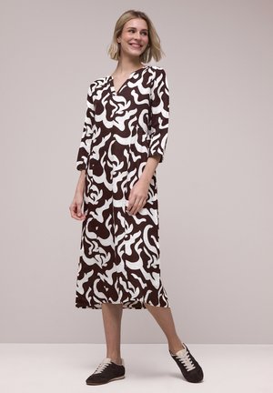 Kvinde iført en brun og hvid abstrakt mønstret midikjole med trekvartærmer, parret med sorte og hvide sneakers, smilende og poserende.