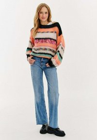 Wielokolorowy sweter z dzianiny w paski w odcieniach różu, pomarańczu i czerni; zestawiony z jasnoniebieskimi dżinsami i czarnymi, masywnymi butami.