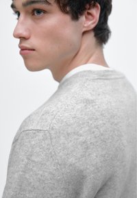 Szary sweter z dzianiny o fakturowanym wykończeniu, z okrągłym dekoltem i widocznymi szwami na ramionach, na neutralnym tle.