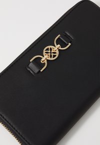 Billetera de cuero negro con herrajes dorados, que presenta un acento de diseño circular en la parte frontal, cierre de cremallera y superficie texturizada.