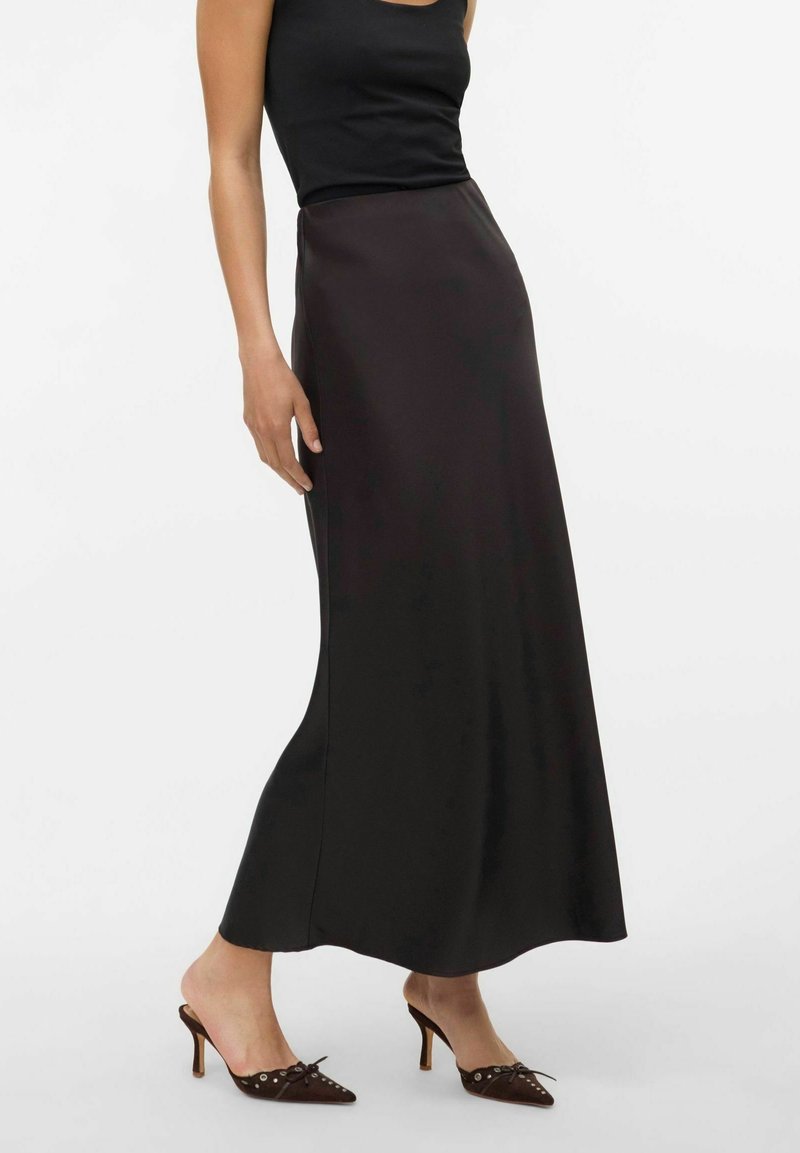 Femme portant une longue jupe noire en satin, un top noir sans manches et des chaussures à talons hauts pointues noires avec un détail de nœud.