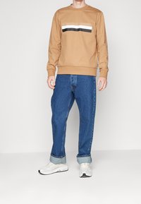 Felpa marrone con una grafica a righe in bianco e nero, abbinata a jeans larghi blu e scarpe da ginnastica bianche. Look semplice e casual.