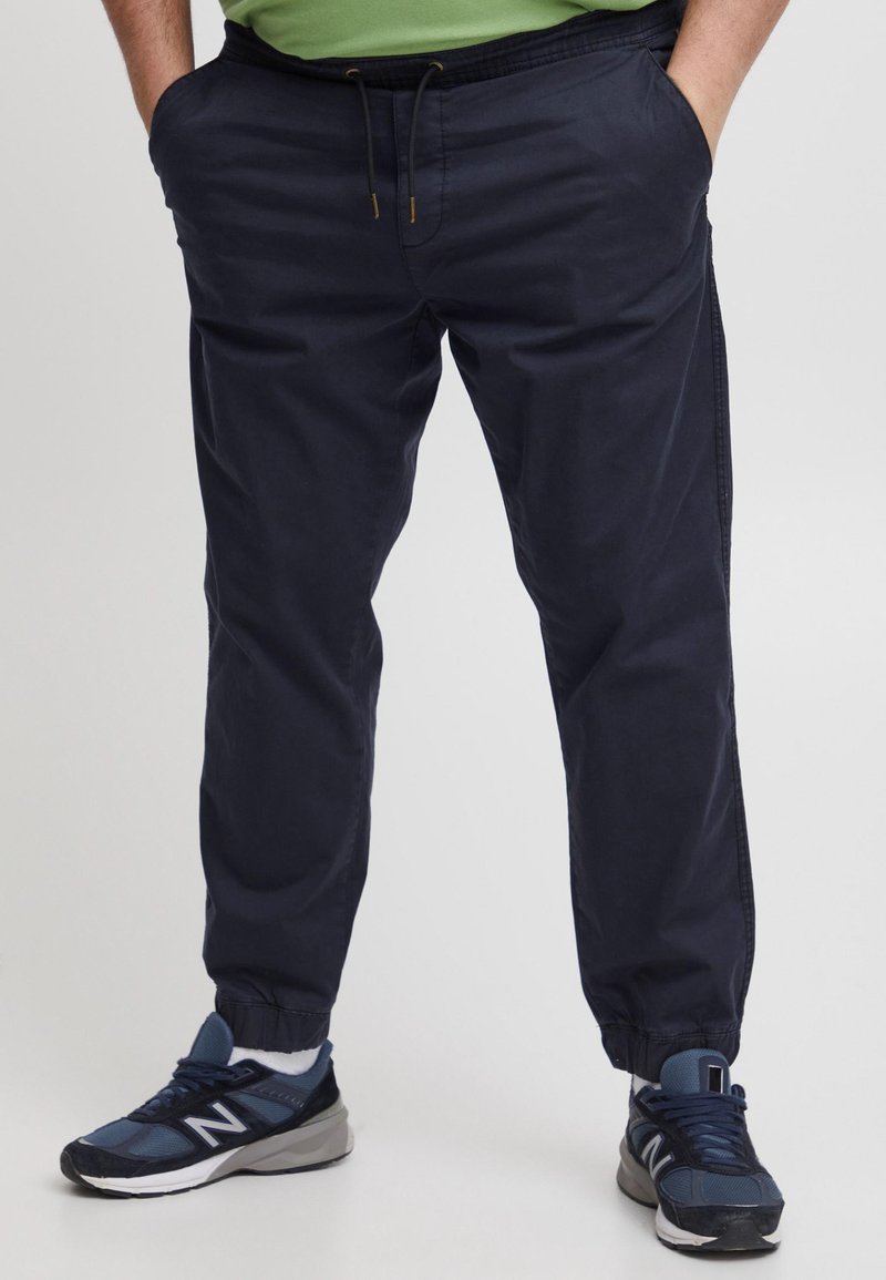 Marineblauwe broek met elastische taille, zijzakken, enkelboorden en een gladde textuur. Gecombineerd met blauwe sportschoenen met zwarte accenten.