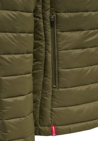 Olivgrüne, isolierte Jacke mit gestepptem Design, mit seitlicher Reißverschlusstasche und rotem Markenlabel unten links.