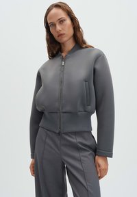 Giubbotto bomber grigio con texture liscia, orlo a costine e chiusura con zip. Presenta una tasca laterale ed è abbinato a pantaloni sartoriali grigi.