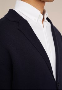 Tmavě modrý pletený kardigan s texturovaným povrchem, s límcem šálu a bílou košilí zapnutou knoflíky pod ním. Čistý design s jemnými detaily.