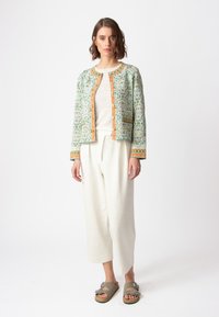 Zelena jakna s uzorkom i narančastim detaljima, uparena s laganom majicom i bijelim širokim hlačama. Model nosi sive sandale s kopčama.