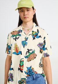 Chemise crème à manches courtes avec des motifs graphiques colorés représentant des plantes, des animaux et des figures. À porter avec un jean bleu et une casquette verte.