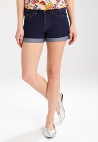 Pantalones cortos de mezclilla con un lavado azul oscuro y dobladillos enrollados. Presentan bolsillos delanteros y cierre de cremallera. Usados con zapatos plateados de tacón bajo.