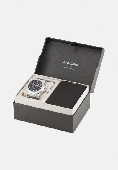 Srebrna ura s temno vzorčasto površino in kovinskim pasom, prikazana v črni škatli z belo notranjostjo ter majhnim brošurjem ob njej.