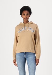 Hoodie beige avec une poche kangourou à l'avant, des cordons de serrage et "GANT 1949" imprimé en bleu. Porté avec un jean bleu clair, offrant une coupe décontractée.