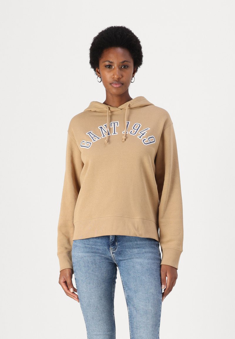 Hoodie beige avec une poche kangourou à l'avant, des cordons de serrage et "GANT 1949" imprimé en bleu. Porté avec un jean bleu clair, offrant une coupe décontractée.