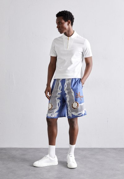 Weißes Poloshirt mit Reißverschlusskragen, kombiniert mit blauen Shorts, die mit aufwendigen Mustern und Akzenten in Grau und Braun verziert sind. Weiße Sneakers runden das Outfit ab.