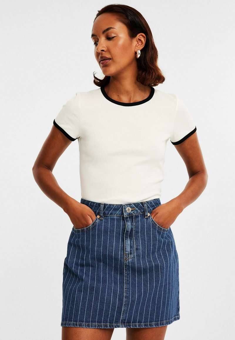 Weißes, geripptes T-Shirt mit schwarzem Saum, kombiniert mit einem Jeansrock mit blauen vertikalen Streifen und seitlichen Taschen. Legerer Schnitt und Design.