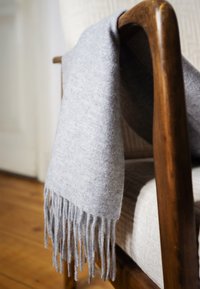 Sciarpa di lana grigio chiaro con frange appoggiata al braccio in legno di una sedia imbottita beige su un pavimento in legno.