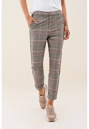 Personne portant un pantalon slim à carreaux gris et roses avec des bandes latérales noires, des baskets beiges et un haut blanc, debout avec une main dans la poche.