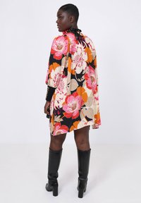Zwarte bloemencape jurk, versierd met grote roze en oranje bloemen, met lange mouwen en een strikdetail bij de hals. Gepaard met zwarte kniehoge laarzen.