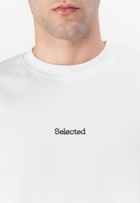 Hvit genser med rund hals og ordet "Selected" brodert i svart på brystet. Glatt stoffstruktur.