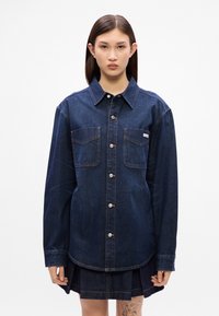 Τζιν πουκ άμισο σε σκούρο μπλε χρώμα με κλασικό γιακά, δύο τσέπες στο στήθος και ασημένια κουμπιά. Διαθέτει μακριά μανίκια και καμπυλωτό στρίφωμα.