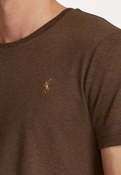 Ruskea puuvillapaita, jossa on pyöreä kaula-aukko ja lyhyet hihat, vasemmassa rinnassa on pieni brodeerattu polo-pelaajan logo.