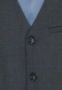 Tkanina szarej marynarki o subtelnym wzorze w kratę w odcieniu niebieskim. Posiada klapę z wcięciem, czarny detal w postaci guzika oraz jasnoniebieską podszewkę.