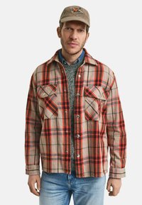 Beige, rot und grün karierte Flanellhemd mit zwei Brusttaschen, Knopfleiste und weicher Textur. Kombiniert mit einem grauen Pullover und blauen Jeans.