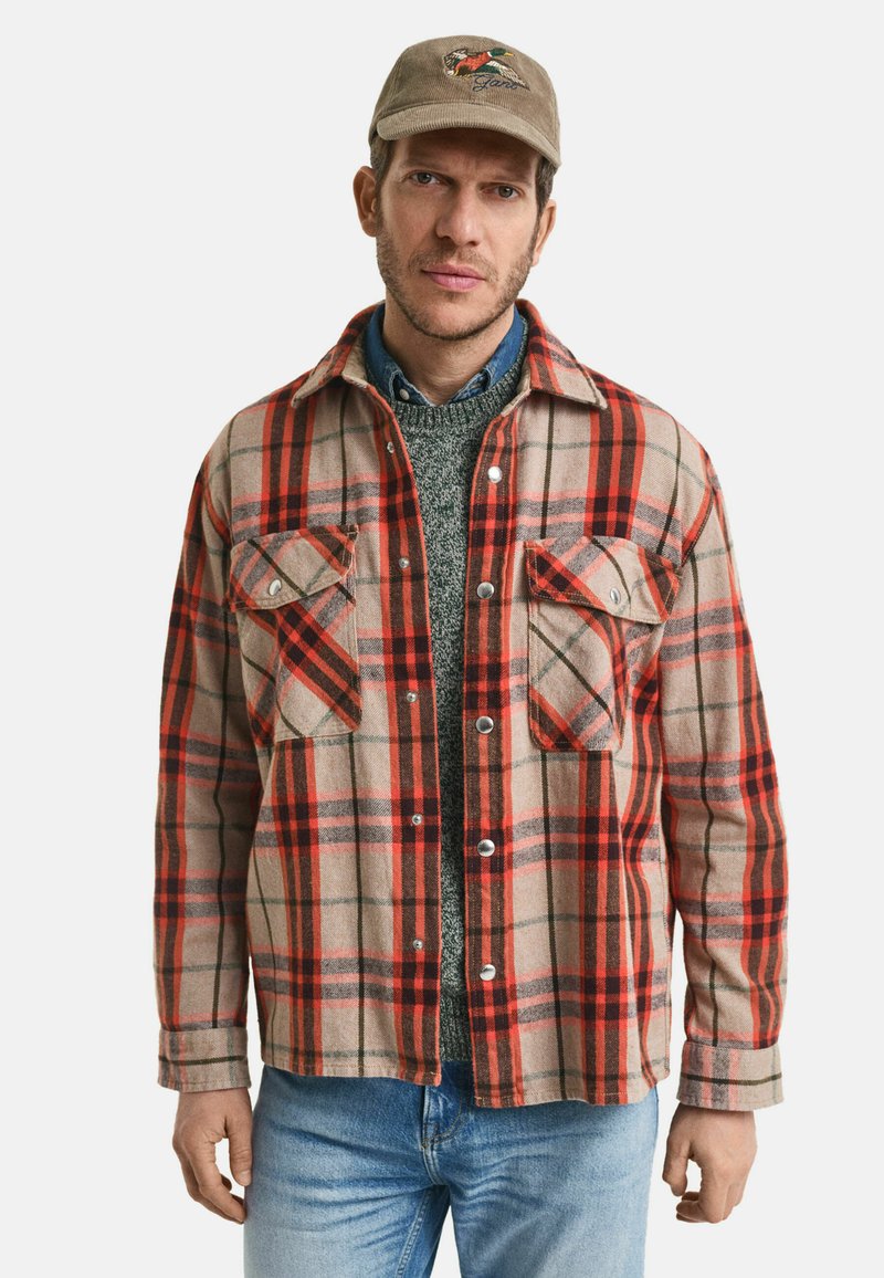 Beige, rot und grün karierte Flanellhemd mit zwei Brusttaschen, Knopfleiste und weicher Textur. Kombiniert mit einem grauen Pullover und blauen Jeans.