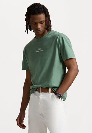 Zaļš kokvilnas T-krekls ar apaļu kaklu un īsām piedurknēm, ar balti izšūtu "Polo Ralph Lauren" logotipu uz krūtīm. Valkāts ar baltiem biksēm.