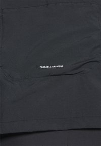 Zwarte opvouwbare kledingstuk met een geperforeerde textuur. Bevat witte tekst met de tekst "OPVOUWBAAR KLEDINGSTUK." Eenvoudig ontwerp zonder aanvullende accenten.