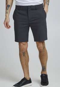 Mørkegrå skreddersydde shorts laget av blandet stoff med knappelukk og to sidelommer, båret med svarte joggesko.