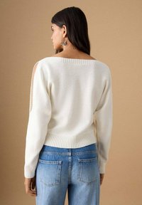 Beli pleteni pulover z širokim izrezom in odprtimi izrezki na ramenih. Rebrasti zaključki na rokavih in spodnjem robu, kombiniran z svetlo modrimi kavbojkami. Gladek tekstura.