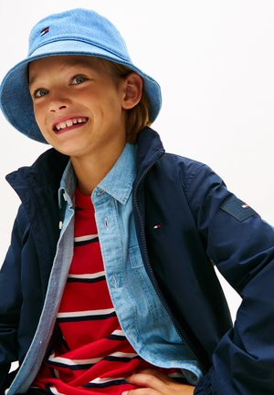 Nasmijano dijete koje nosi svijetloplatu kapu na kant, tamnoplavu jaknu, svijetlu traper košulju i crvenu majicu s prugama, pozira na bijeloj pozadini.