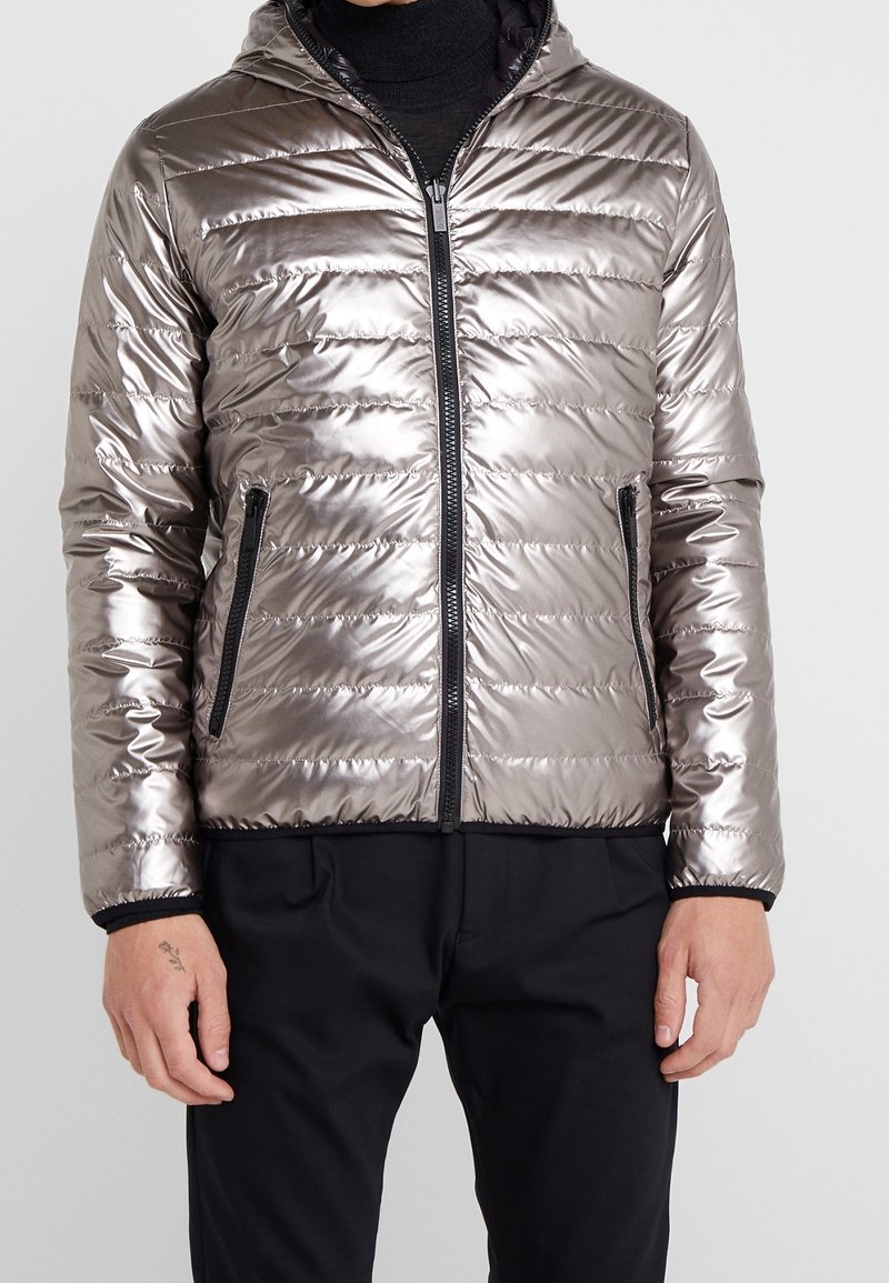 Mann trägt eine metallisch silberne Steppjacke mit Frontreißverschluss und Reißverschlusstaschen an den Seiten, kombiniert mit schwarzen Hosen und einem schwarzen Rollkragenpullover.