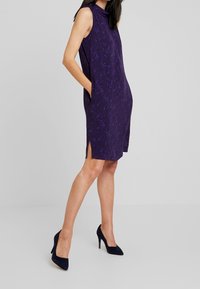 Fioletowa sukienka bez rękawów z dekoltem w łódkę, bocznymi kieszeniami i rozcięciami u dołu; wykonana z teksturowanej tkaniny o subtelnym wzorze.