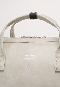 Sac à main Anello gris clair avec deux poignées et une fermeture éclair argentée centrée, présentant un petit logo imprimé et un texte sur le panneau avant.