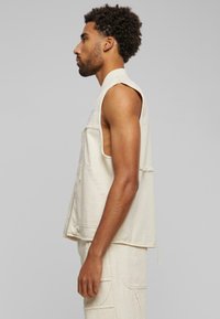 Uten ermer kremfarget denimvest med frontlommer, rå kant og synlige sømdetaljer, vist på en modell i profil.