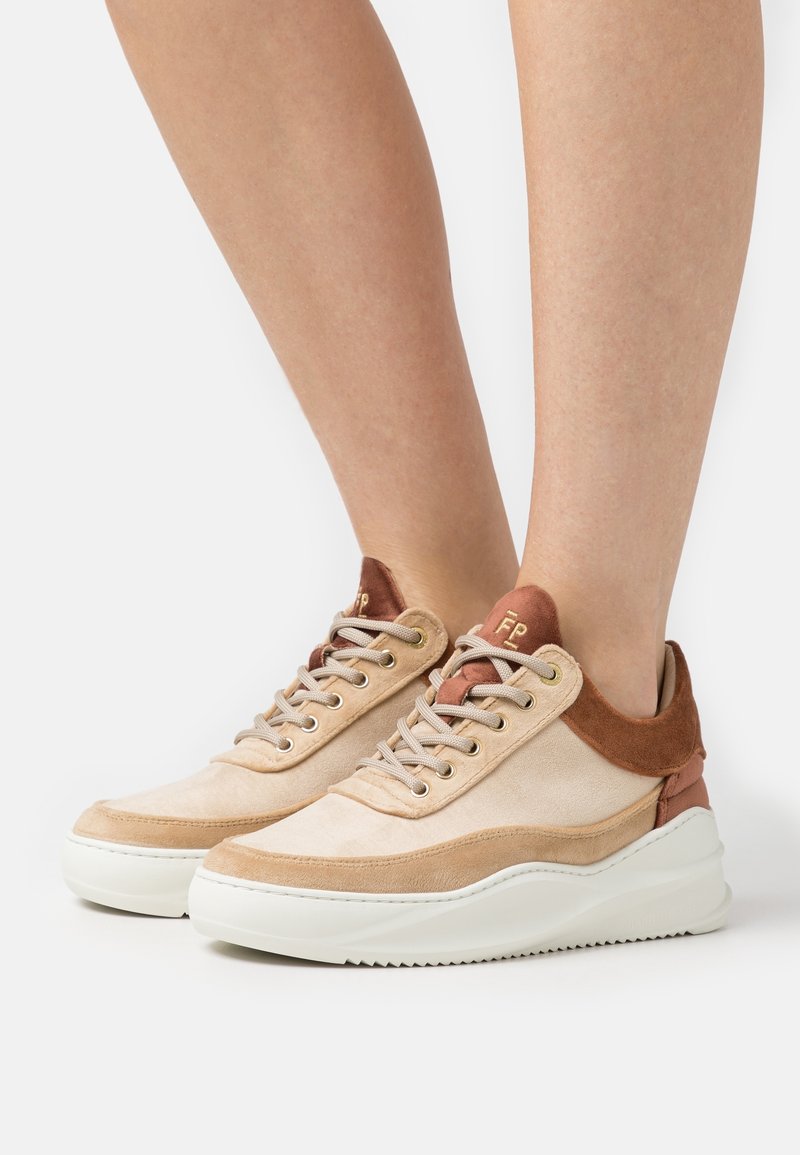 Pieds portant des baskets en daim beige et marron à lacets avec des semelles blanches épaisses sur un fond blanc.