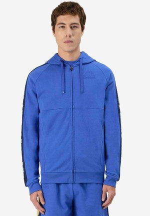 Jeune homme debout portant un sweat à capuche bleu zippé avec un logo sur la poitrine et des manches rayées, assorti à un pantalon bleu assorti.