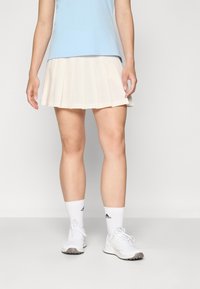 Šviesiai smėlio spalvos raukšlėtas teniso sijonas, derinamas su mėlynu viršumi. Modelis dėvi baltus sportinius batelius ir kojines su juodais logotipo akcentais.