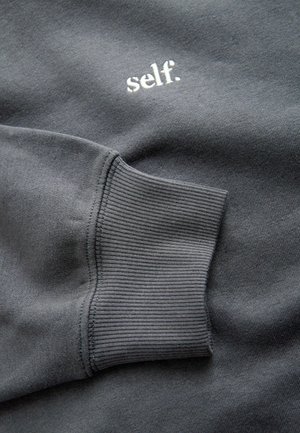 Pelēks džemperis ar ribotām aprocēm; uz tā ar baltu izšūts teksts "self." Mīksta tekstūra, ikdienišķs dizains, minimālistisks izskats.