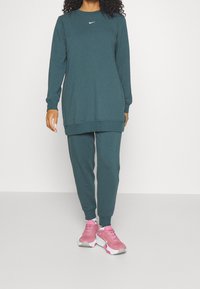 Zelená mikina s kulatým výstřihem a dlouhými rukávy, doplněná o odpovídající kalhoty; obsahuje bílé logo Nike a růžové sportovní boty.
